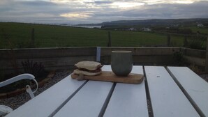 Ein Picknicktisch mit einem Sandwich und einer Keramiktasse, mit Blick auf ein Wiesenfeld und entfernte Häuser unter bewölktem Himmel.