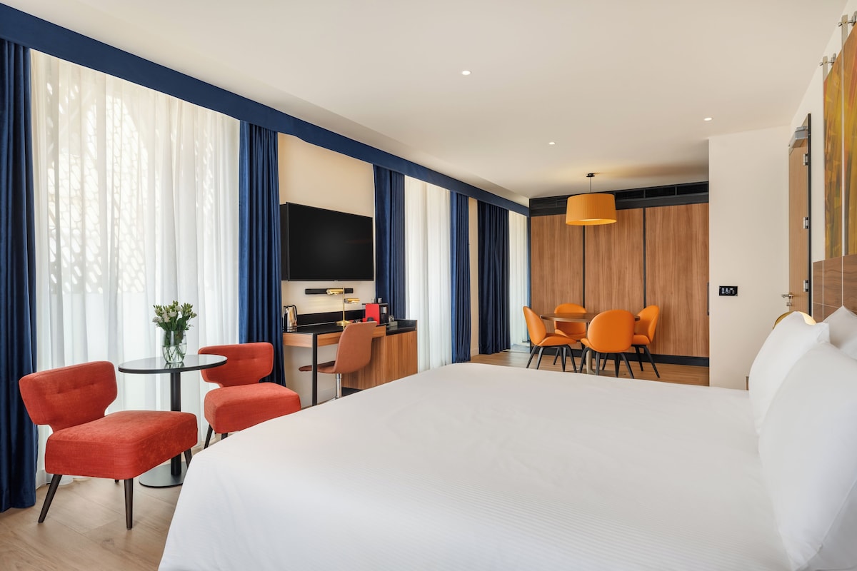 Quarto de hotel com uma cama grande, duas cadeiras vermelhas, uma pequena mesa com um vaso, uma escrivaninha com cadeira e uma televisão.