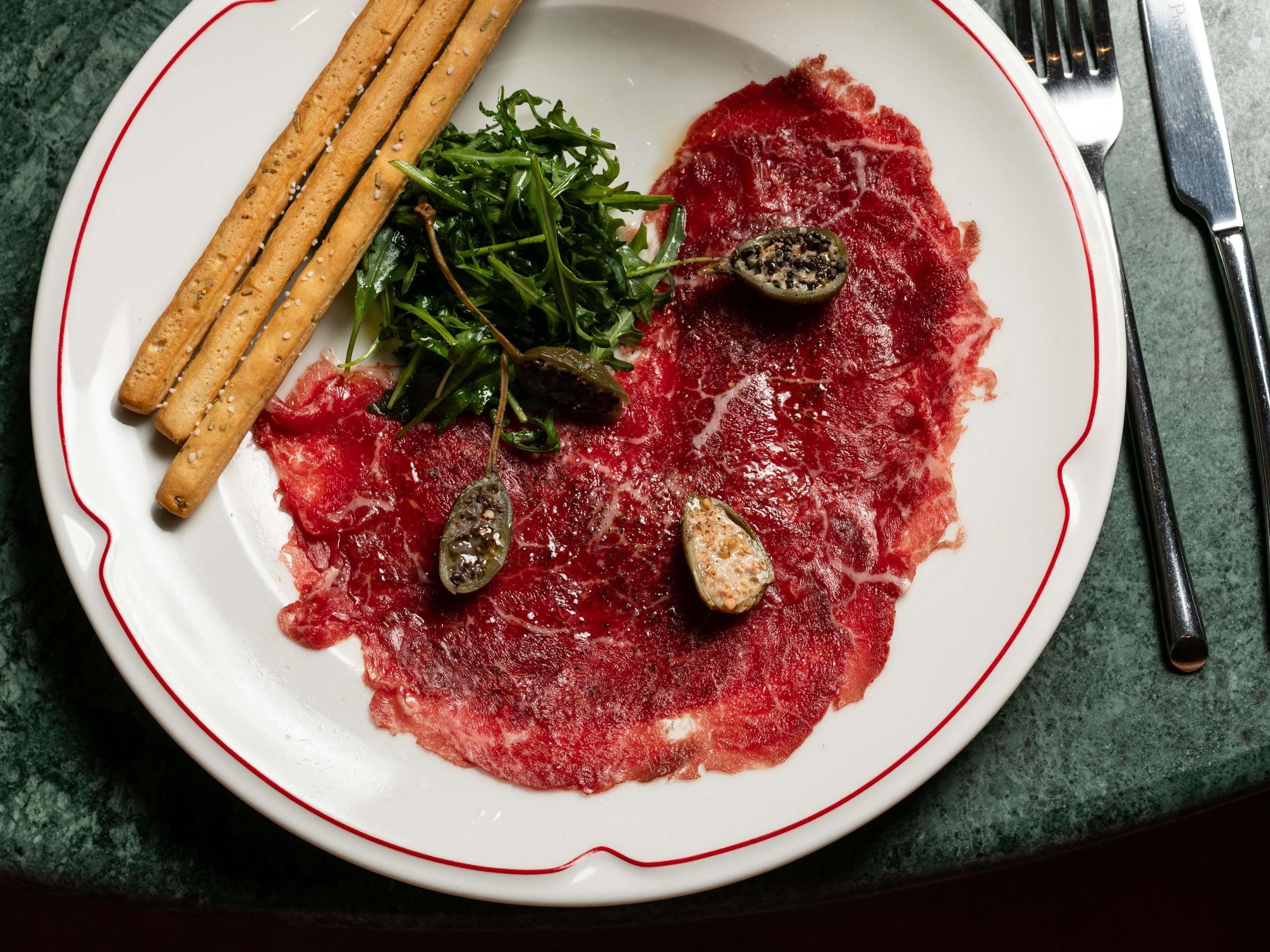 A plate with raw beef, greens, and pickled items on a white plate with a red rim.