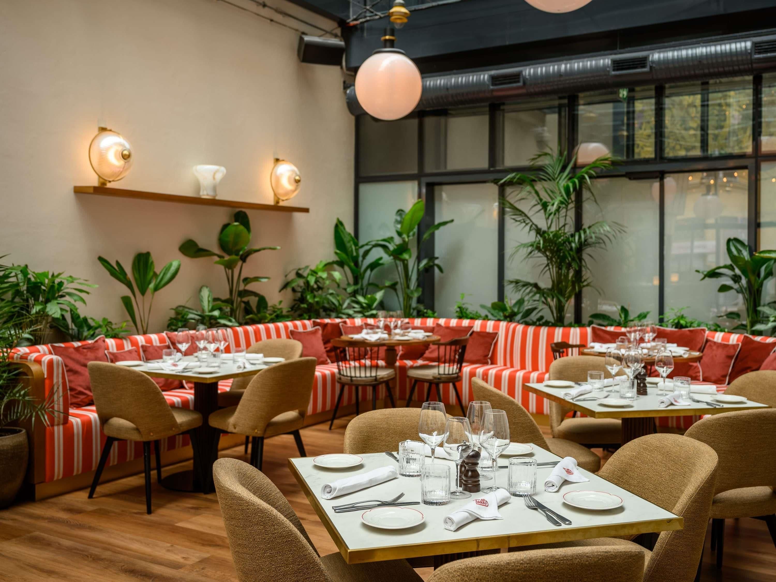 A restaurant with a cozy seating area, tables set with plates and glasses, and a large window with plants outside.