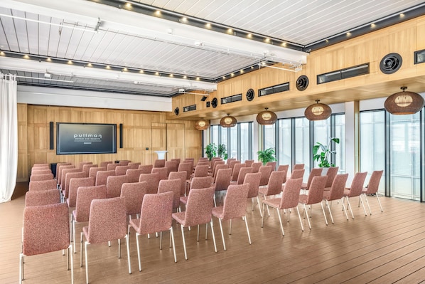 Uma sala de conferências com fileiras de cadeiras, uma tela grande e painéis de madeira.