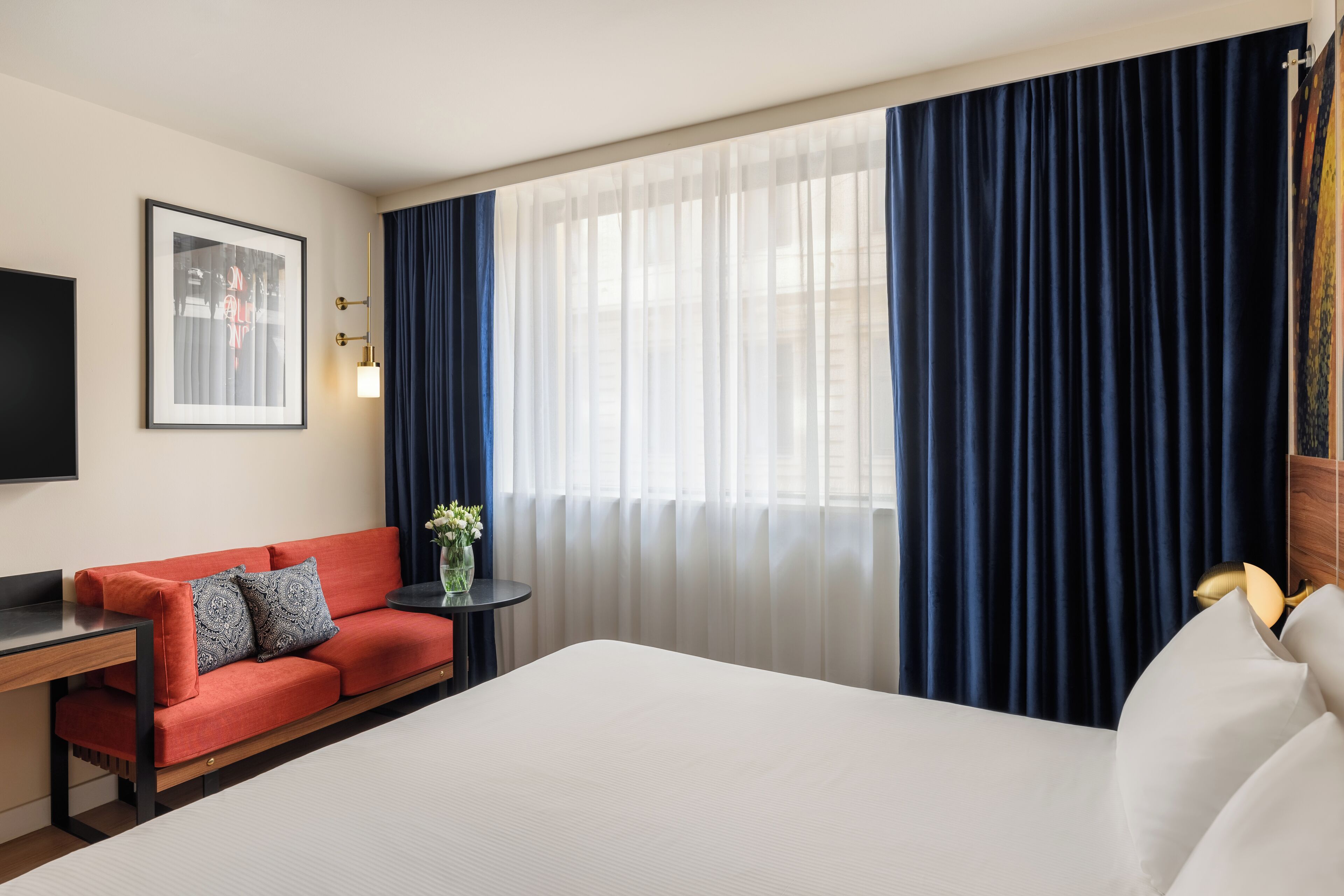 Habitación de hotel con una cama grande, un sofá rojo, una mesita con un jarrón y un ventanal con cortinas.