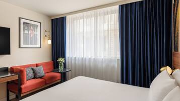 Habitación de hotel con una cama grande, un sofá rojo, una mesita con un jarrón y un ventanal con cortinas.