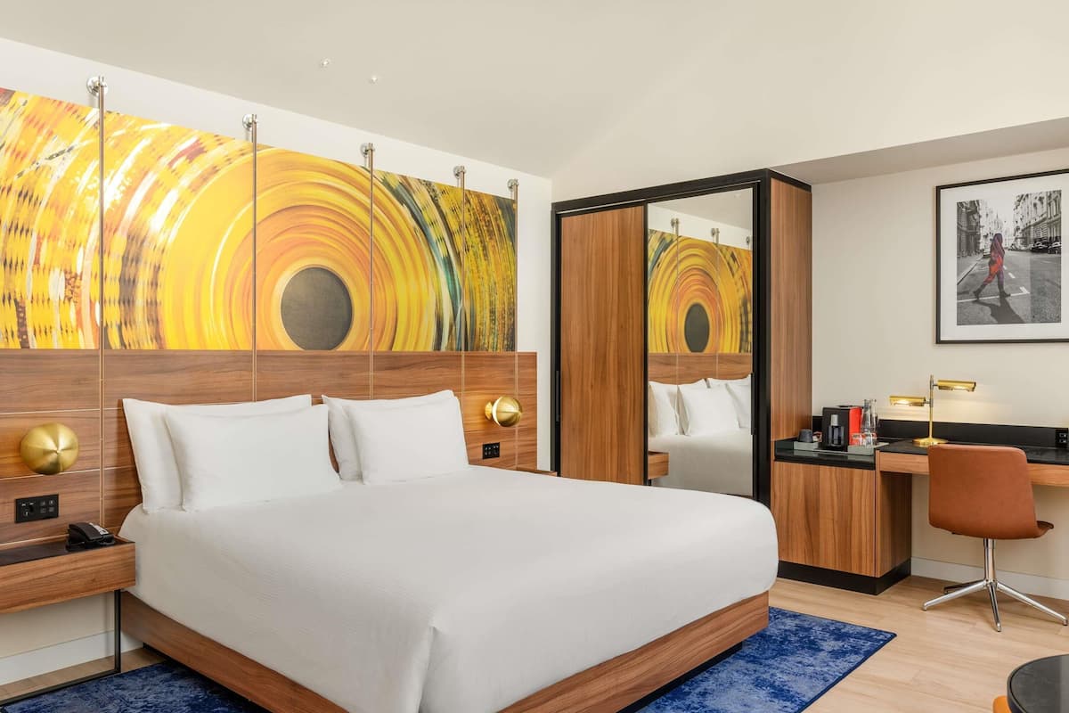 A hotel room with a large bed, a desk, a chair, a mirror, and a painting on the wall.