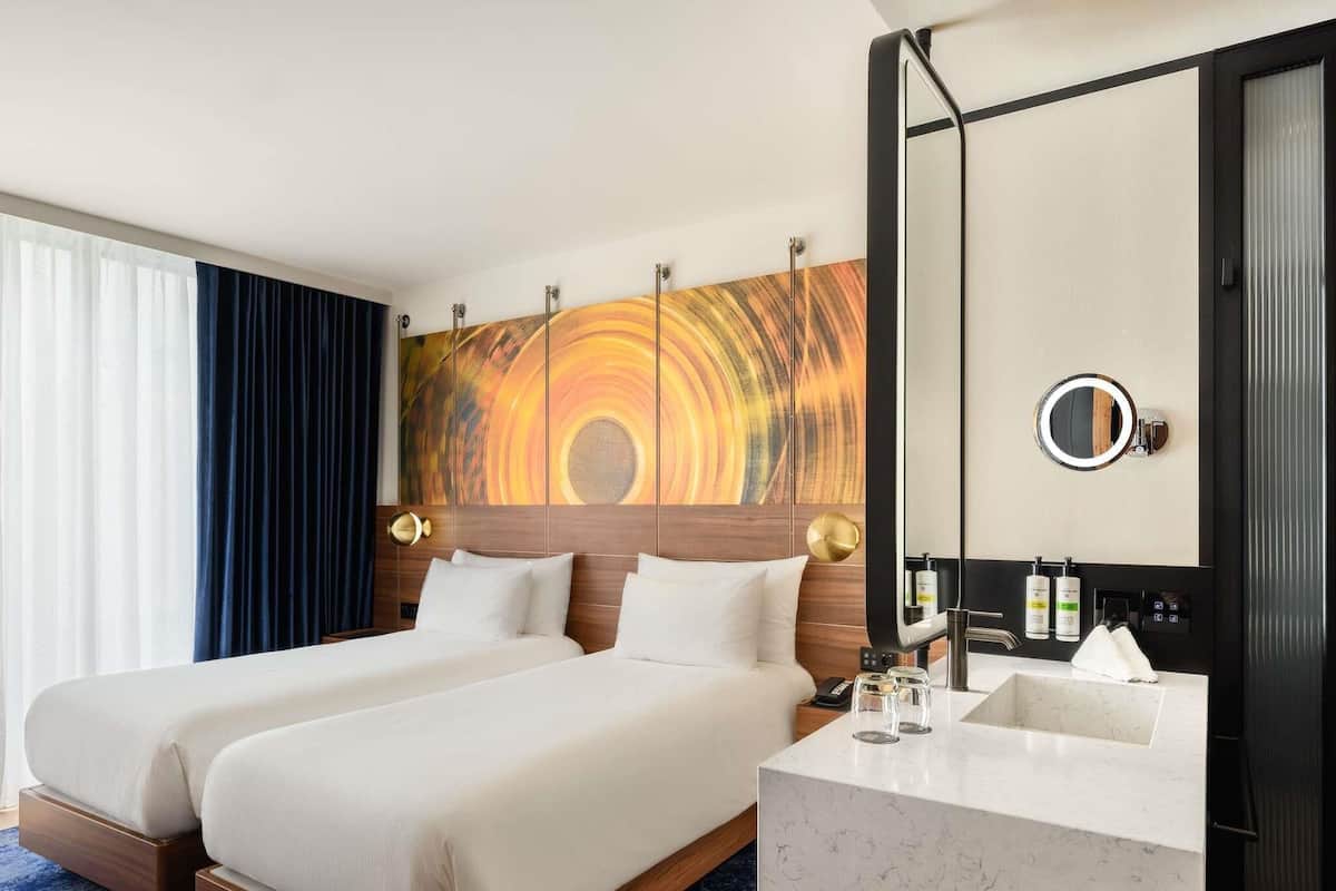 Habitación de hotel con una cama grande, un baño con lavamanos y espejo, y una pared con un cuadro abstracto de colores.
