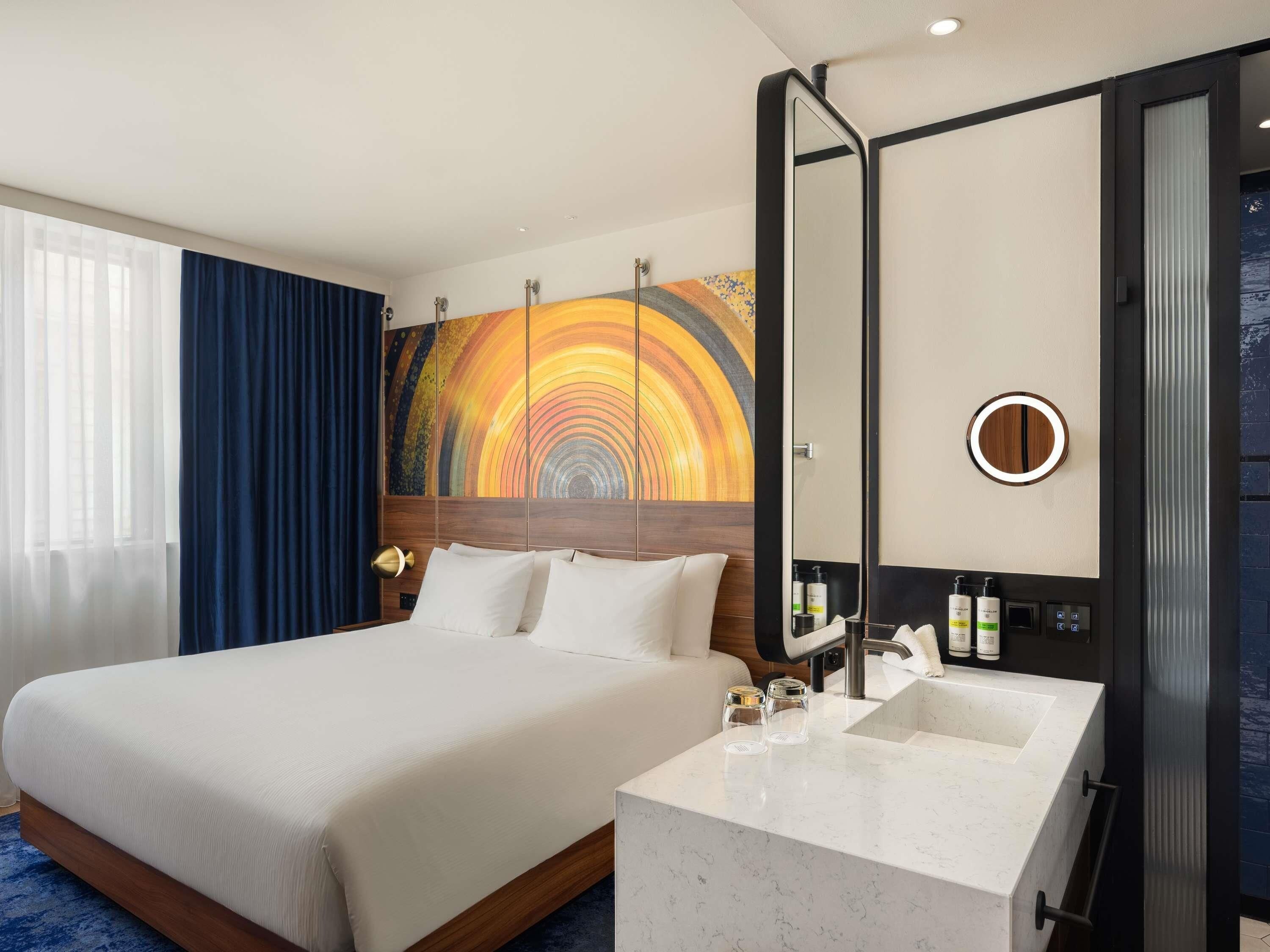 A hotel room with a large bed, a bathroom with a mirror and sink, and a colorful mural on the wall.
