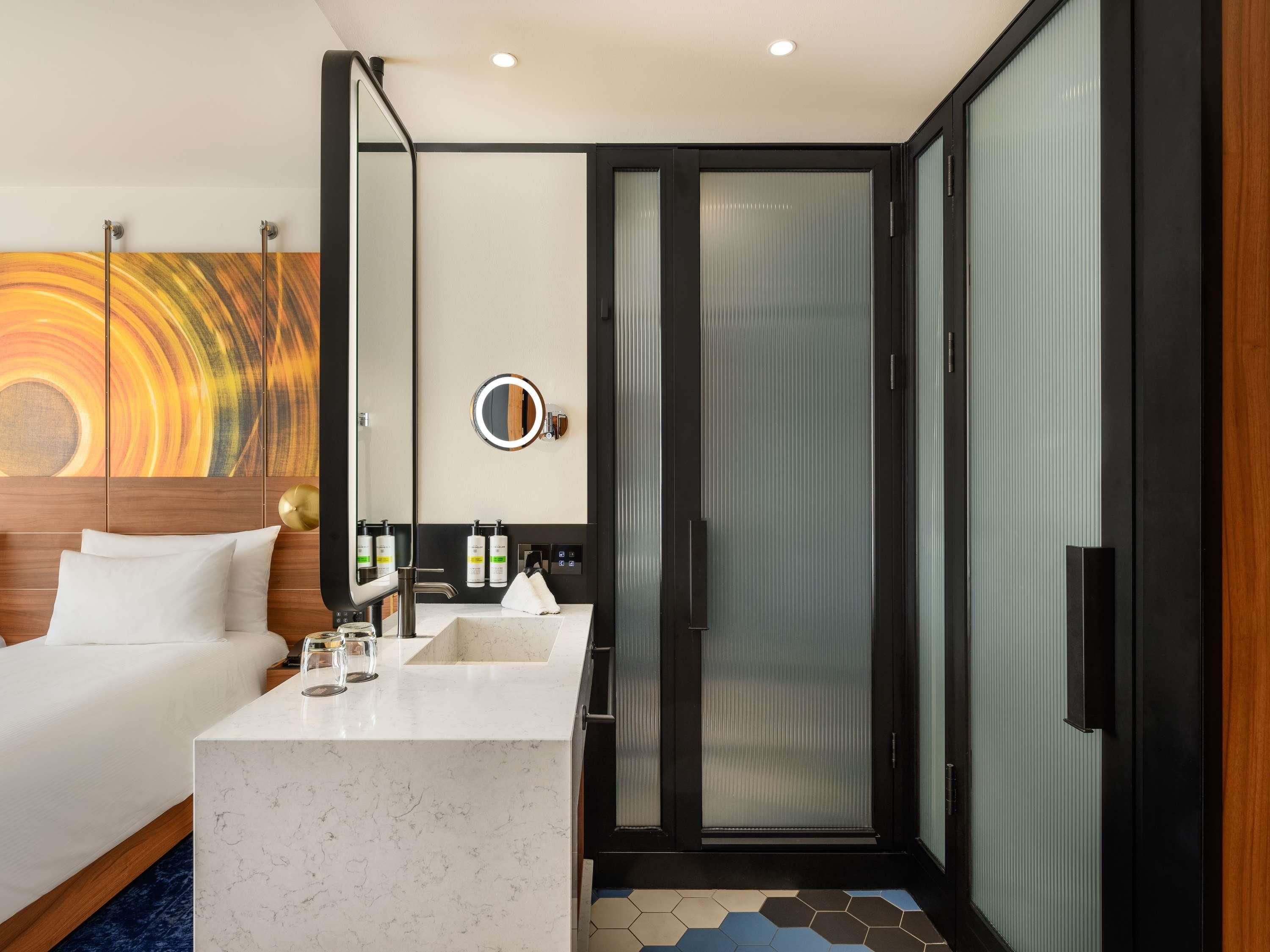 A modern hotel room with a bed, a marble bathroom vanity, a round mirror, and a sliding glass door leading to a shower area.