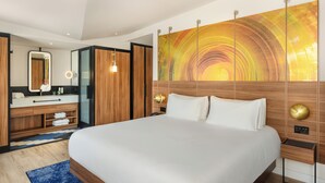 A modern hotel room with a large bed, wooden headboard, a large mirror, a vanity with a sink, and a colorful abstract painting on the wall. - Pullman Budapest (Budapest)