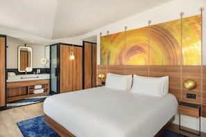 Una habitación de hotel moderna con una cama grande, cabecera de madera, un espejo amplio, tocador con lavamanos y un cuadro abstracto colorido en la pared.