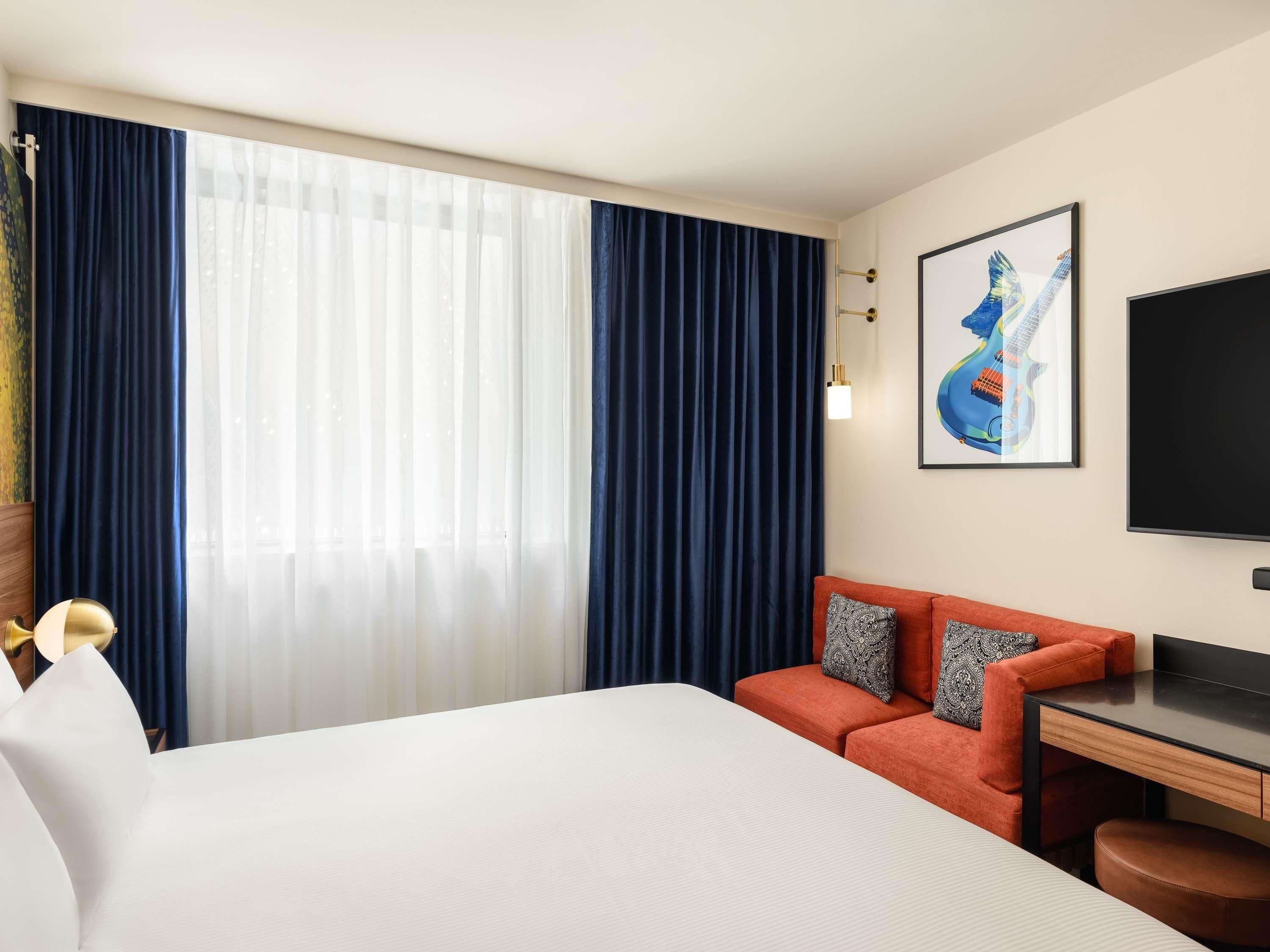 A hotel room with a bed, a desk, a chair, a TV, and a painting on the wall.