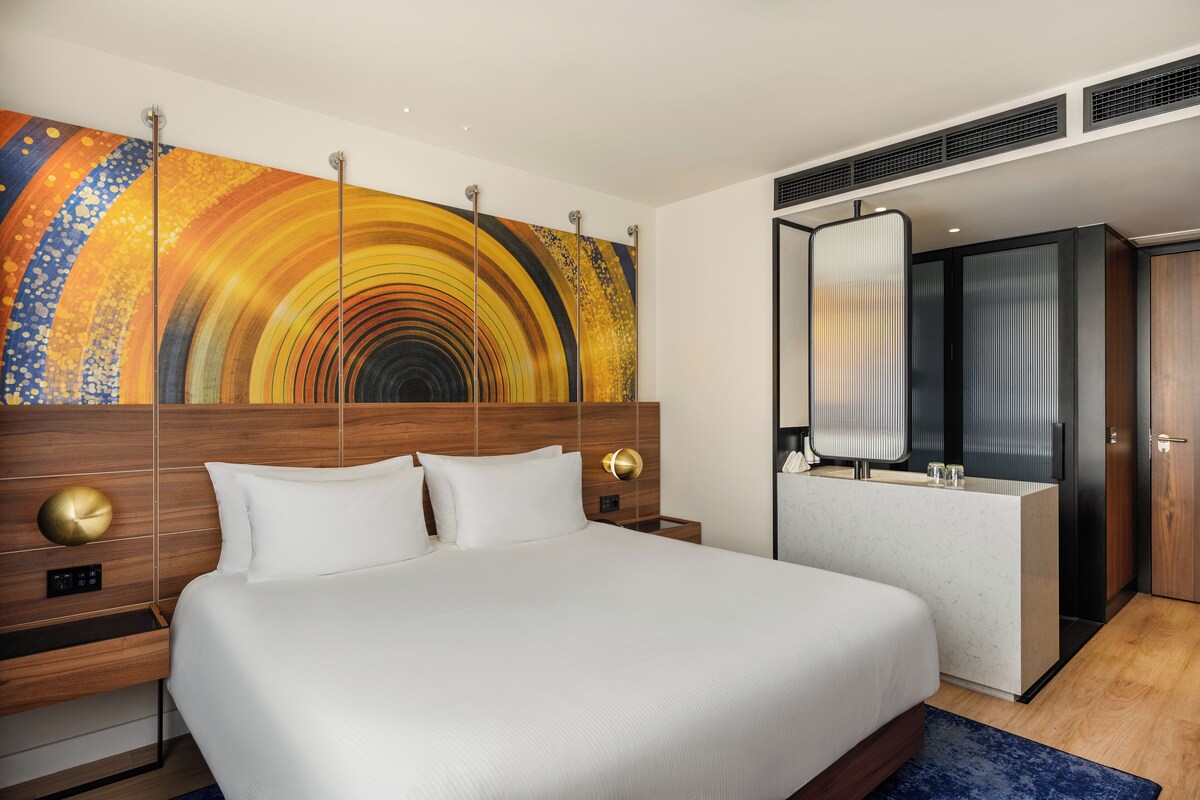 Quarto de hotel moderno com cama grande, cabeceira de madeira, uma obra de arte na parede, banheiro com espelho e uma pequena mesa de trabalho.