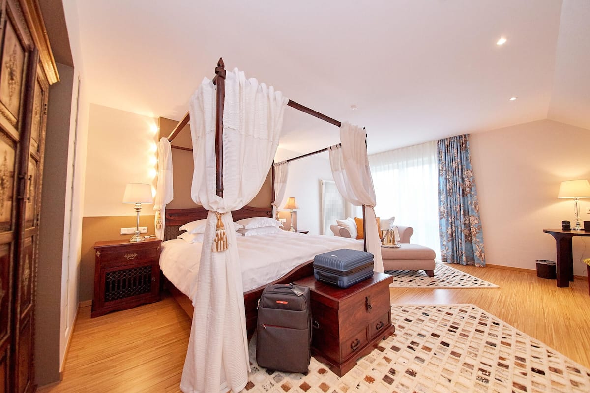 A four-poster bed with white bedding, a bedside table with a lamp, a suitcase on a chest of drawers, and a patterned rug on the floor.