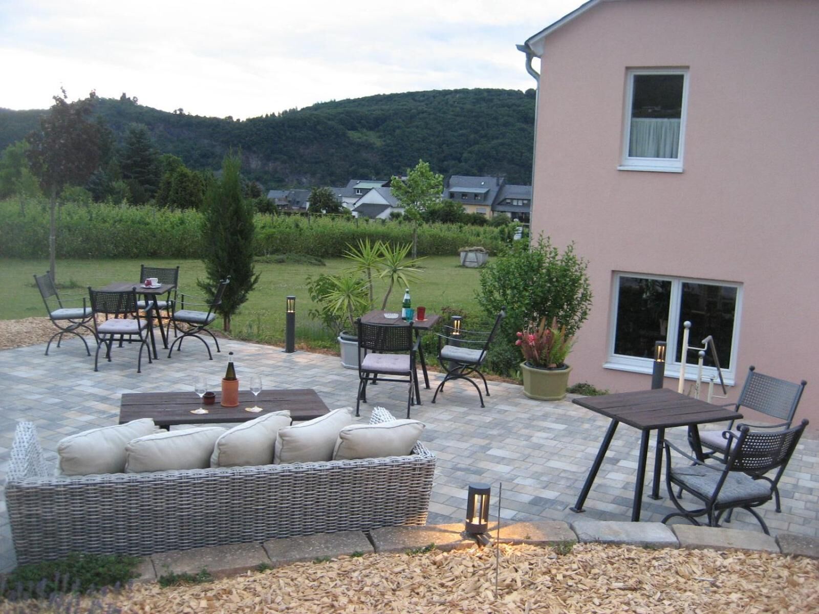 Eine Terrasse im Freien mit einem Korbsessel, einem Esstisch und Stühlen, umgeben von Blumentöpfen und Blick auf eine Hügelkuppe.