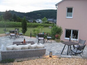 An outdoor patio with a wicker sofa, dining table, and chairs, surrounded by potted plants and a view of a hillside. - GästeHaus Auler Hof (Kröv)