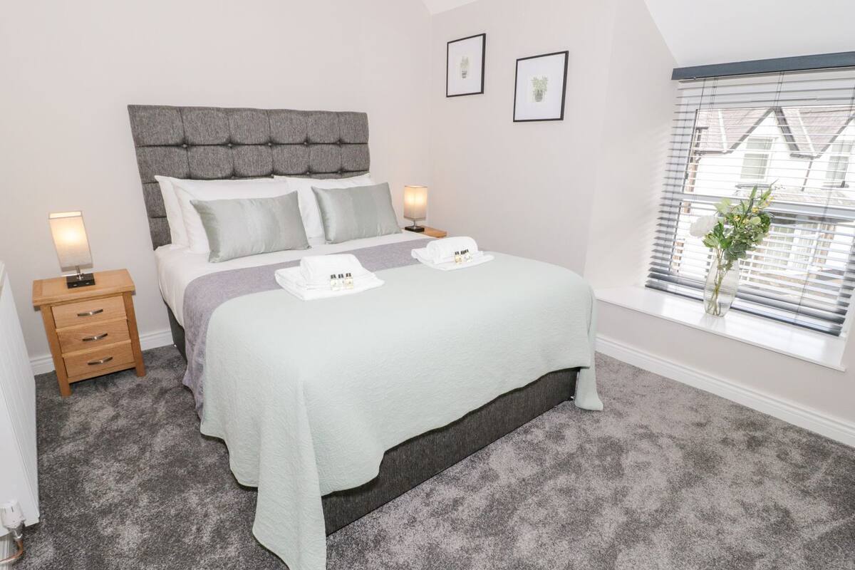 A neatly arranged bedroom with a bed, bedside tables, a window with blinds, and framed pictures on the wall.