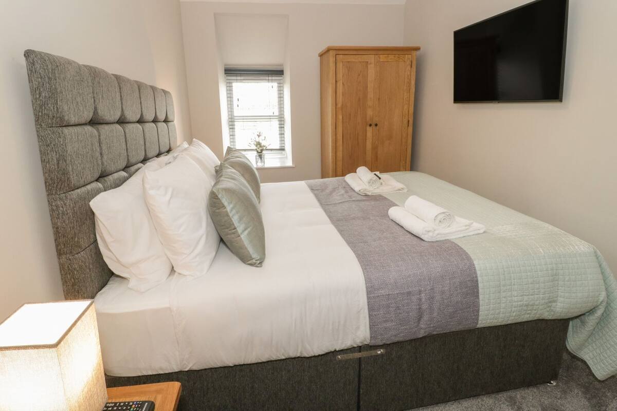 A neatly made bed with pillows, a blanket, and rolled towels. There is a wooden wardrobe, a television on the wall, and a window with blinds.