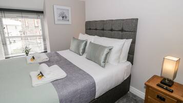 A neatly made bed with a grey headboard, white bedding, and a grey throw blanket. There is a bedside table with a lamp and a remote control. A framed picture hangs on the wall above the bed.