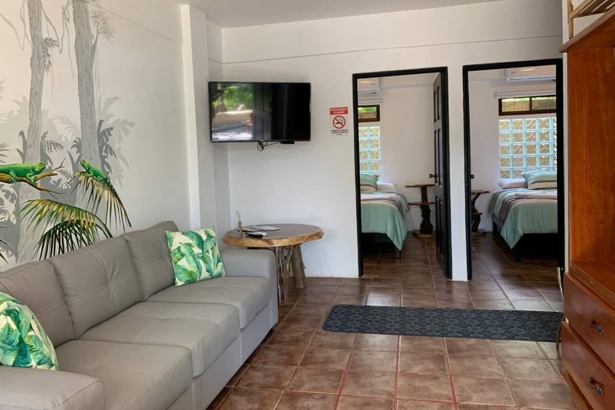 Una habitación con sofá, una mesita redonda, televisor montado en la pared y una cama con colcha verde.