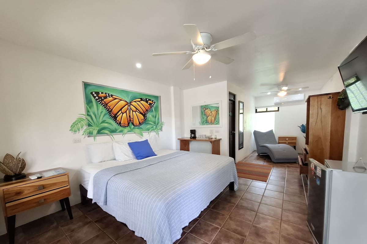 Una habitación de hotel con cama, escritorio, silla, ventilador de techo y refrigerador.