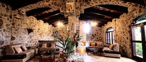 A stone-walled room with a high ceiling, furnished with sofas and a coffee table, and a large window providing natural light.
