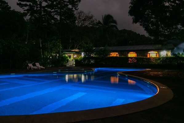 A nighttime swimming pool with a curved edge, surrounded by lush greenery and a building with illuminated windows. - Malekus Mountain Lodge (Guayabo)