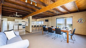 Een ruime woonkamer met houten plafond, een eettafel met stoelen, een keuken op de achtergrond en een groot raam met uitzicht.