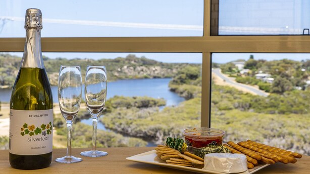 Een fles Churchview Silverleaf sparkling brut wijn, twee glazen, een bord met crackers, kaas en een klein schaaltje jam, geplaatst op een tafel met uitzicht op een meer en groen.