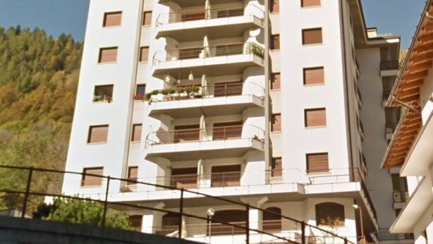 Un hotel de varios pisos con balcones, techo plano y montañas al fondo.