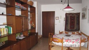 Un comedor con una mesa redonda, sillas y una lámpara colgante. Se ve una estantería con libros y una puerta al fondo.