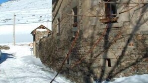A stone building with a sloped roof, a small attached structure, and a snow-covered mountain in the background. - Ker Carlines (Targassonne)