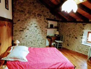 Une pièce avec un mur en pierre, un lit recouvert d’une couverture rose, un bureau en bois, une chaise et une lampe.