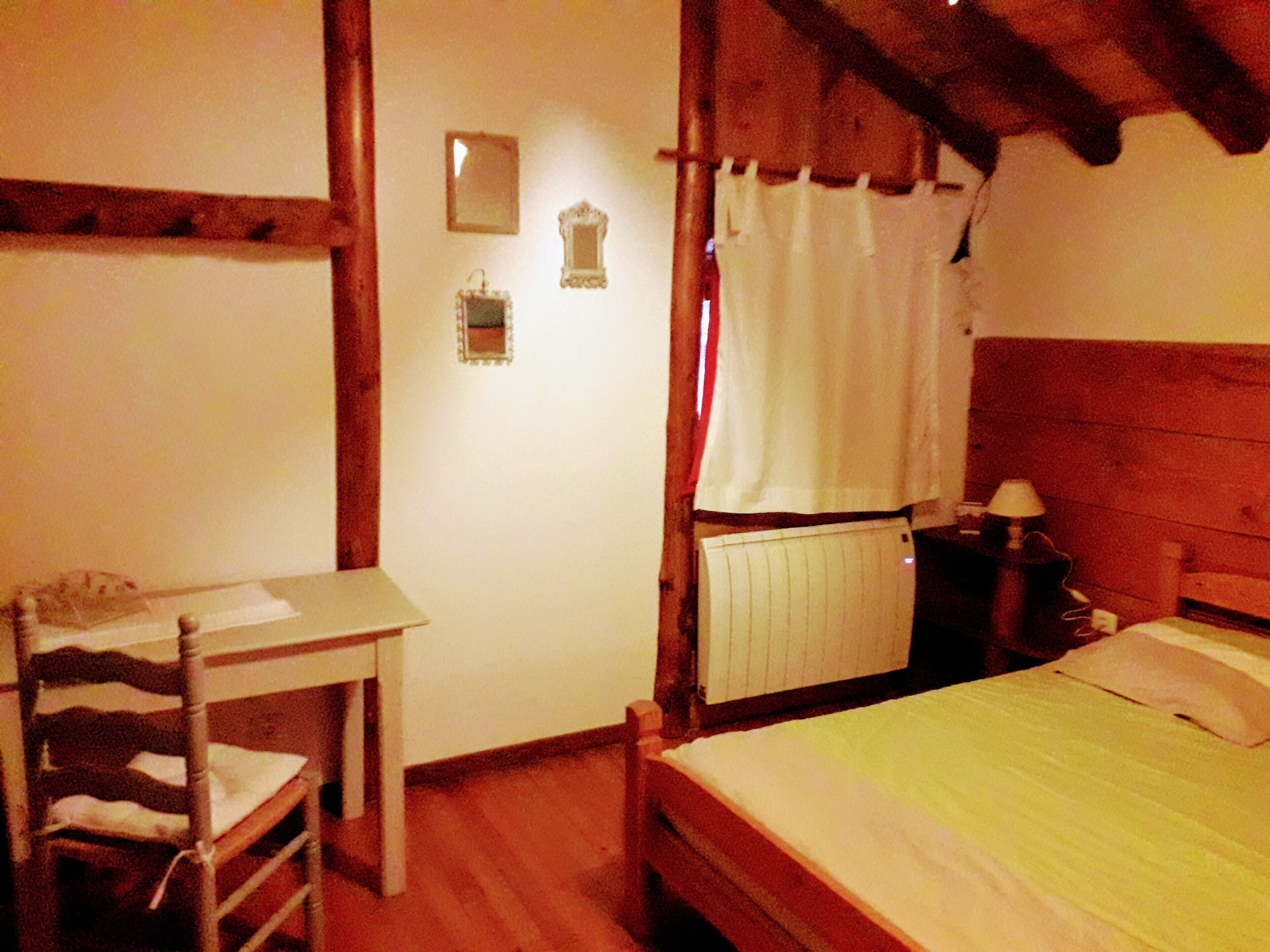 A bedroom with a wooden bed, a small table, a chair, a radiator, and a mirror on the wall.