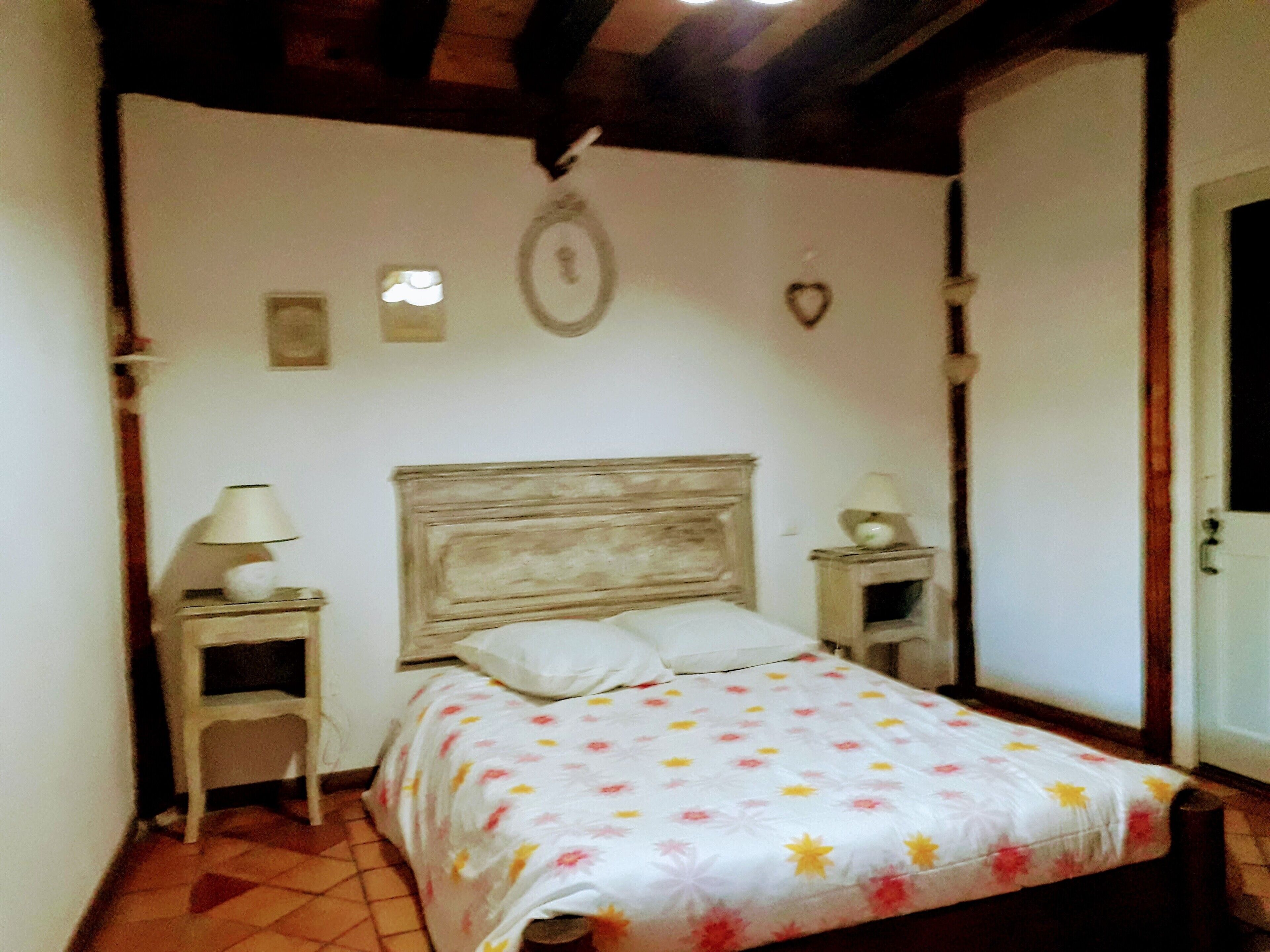 Une chambre à coucher avec une tête de lit en bois, une couvre-lit à motifs floraux, deux tables de chevet avec des lampes et une petite décoration murale.