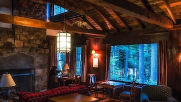 Un intérieur chaleureux de chalet avec une cheminée en pierre, des poutres en bois, une grande fenêtre donnant sur la forêt et un canapé à carreaux rouges.