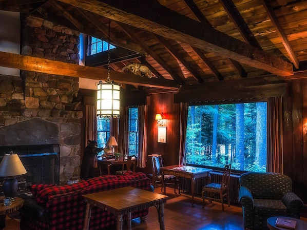 A cozy lodge interior with a stone fireplace, wooden beams, a large window with a forest view, and a red plaid sofa.