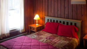 Een slaapkamer met een houten wand, een bed met quilt, een nachtkastje met lamp en een raam met gordijnen.