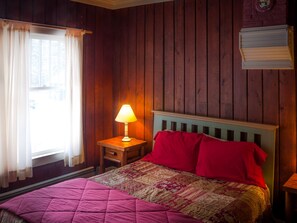 A bedroom with a wooden wall, a bed with a quilt, a nightstand with a lamp, and a window with curtains.
