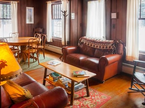 Un salon chaleureux avec un canapé en cuir, une table basse en bois sur laquelle sont posés des livres, une table à manger avec des chaises et de grandes fenêtres ornées de rideaux.