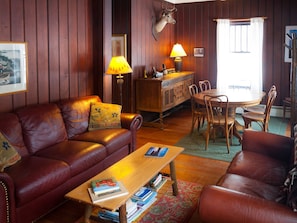 A cozy living room with leather sofas, a wooden coffee table with books, a lamp, and a deer head mounted on the wall. - Gate Cottage at Pet Friendly (Paul Smiths)