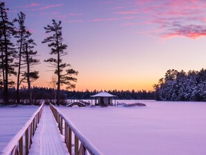 Sentiero innevato che conduce a una piccola costruzione in legno con tetto, circondata da alberi alti e un lago ghiacciato.