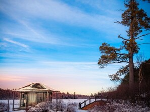 A small, rustic cabin with a glass door, surrounded by snow-covered trees and a clear sky.