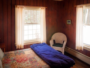 Une pièce aux murs en bois, un lit recouvert d’une couvre-lit à motifs floraux, une chaise et deux fenêtres avec des rideaux.