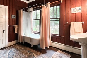 A bathroom with a bathtub, a window with curtains, a sink, and a wooden floor.