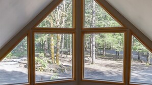 Habitación con techo triangular y ventanas amplias que ofrecen vista a un área boscosa con un sendero de tierra y bancos.