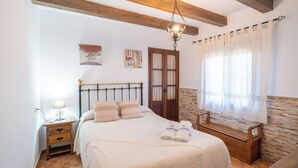 A bedroom with a bed, bedside table, lamp, artwork, wooden bench, and a window with curtains. - Las Adelfas del Gastor by Ruralidays (Cádiz)