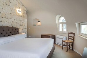 A bedroom with a stone wall, a bed, a chair, a nightstand, a lamp, and a window.