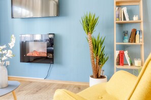 A living room with a flat-screen TV mounted on the wall, a bookshelf with books and a candle, a potted plant, and a yellow sofa. - Scandinavian - pool - fireplace - Trendy (Winterberg)