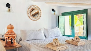 Ein Schlafzimmer mit einem Bett, Kissen, einem Nachttisch mit Lampe, einem Spiegel und Blick ins Freie durch eine grüne Tür.