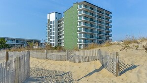 Un hôtel en bord de mer, avec plusieurs étages, une plage de sable en premier plan et une clôture en bois.