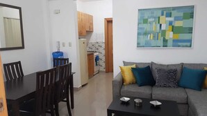 Una sala de estar con un sofá gris, una mesa de centro y un comedor con mesa y sillas. Se ve una cocina al fondo con armarios de madera y salpicadero de azulejos.